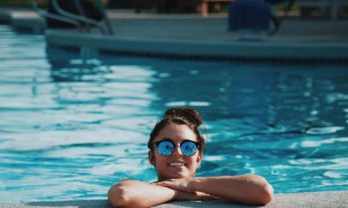 woman with sunglasses, in a pool, rests her arms on the ledge and smiles